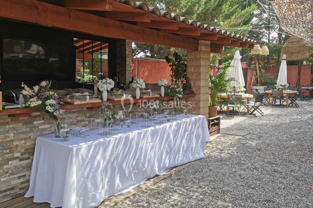 Table dressée avec une nappe blanche devant un comptoir en pierre, dans un espace extérieur décoré de fleurs et de…