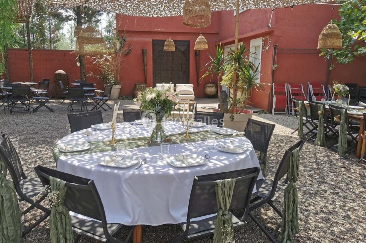 Table ronde dressée avec une nappe blanche, entourée de chaises décorées, dans une cour extérieure ombragée.