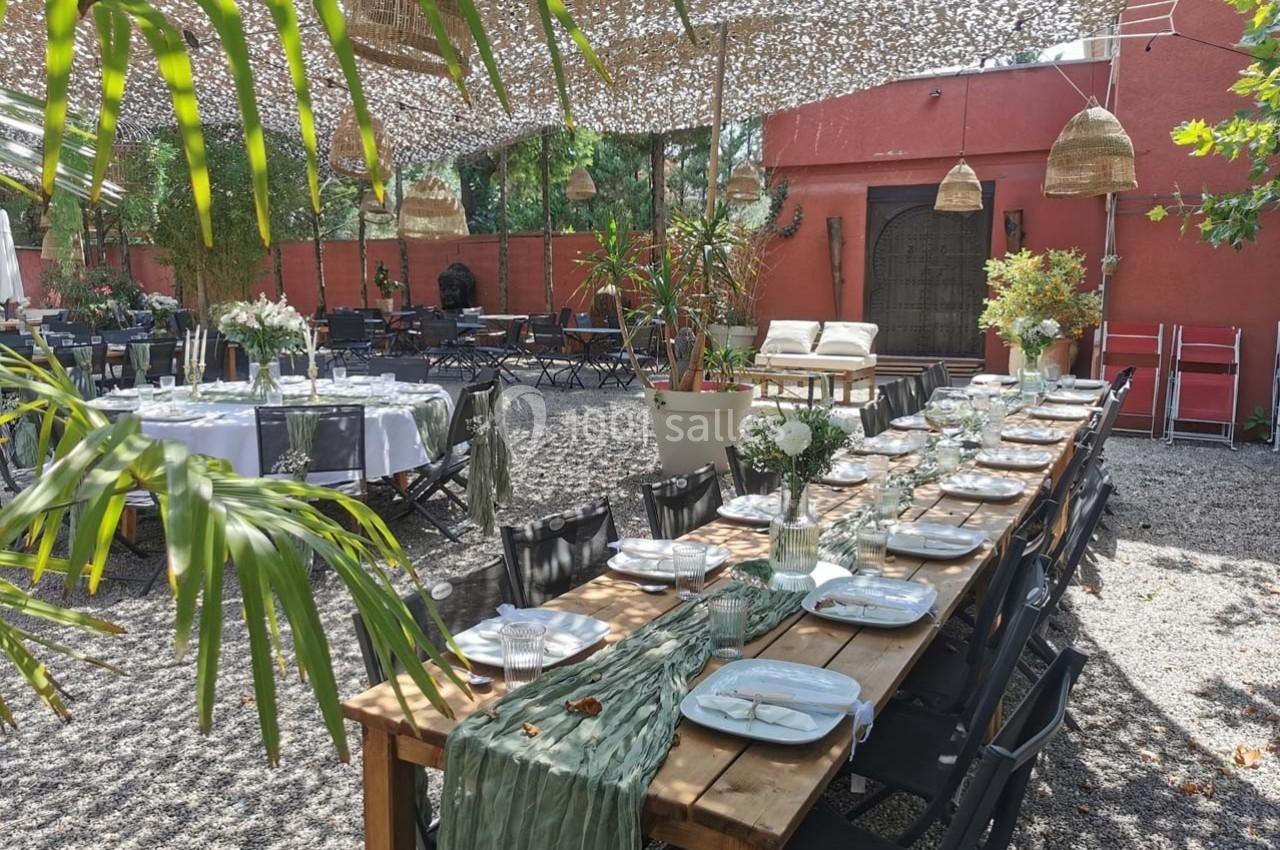 Grande table en bois dressée pour un repas en extérieur, entourée de chaises noires, sous des voiles d'ombrage.
