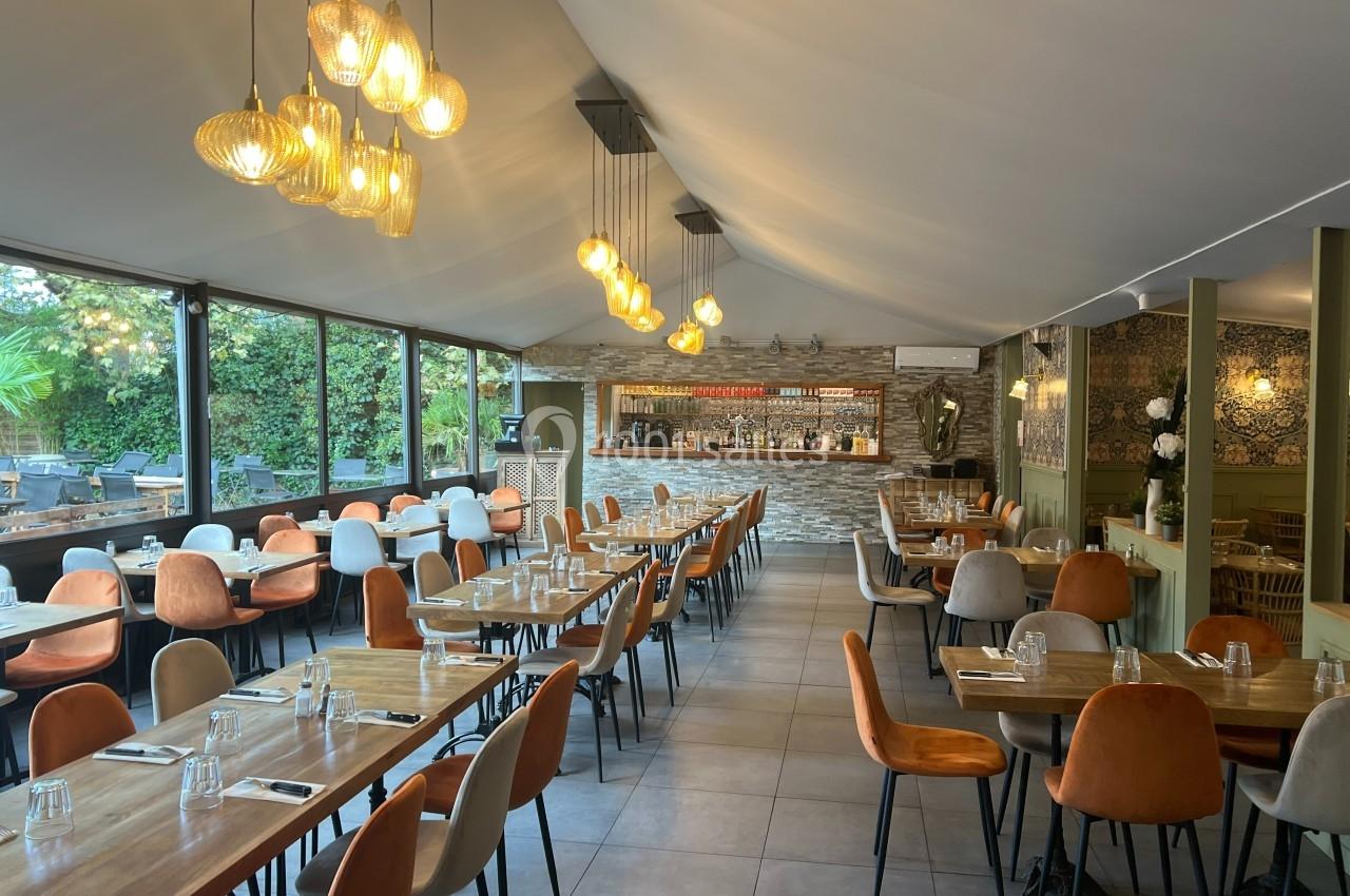 Salle de restaurant lumineuse avec tables en bois, chaises colorées et bar en pierre au fond, éclairée par des suspensions…