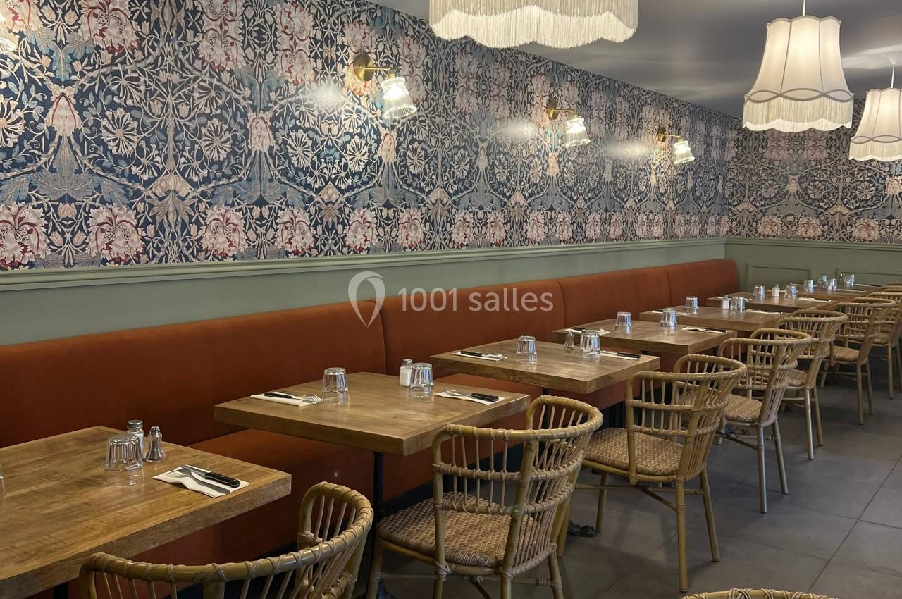 Salle de restaurant avec tables en bois, chaises en rotin, banquette orange et murs ornés d’un papier peint fleuri.