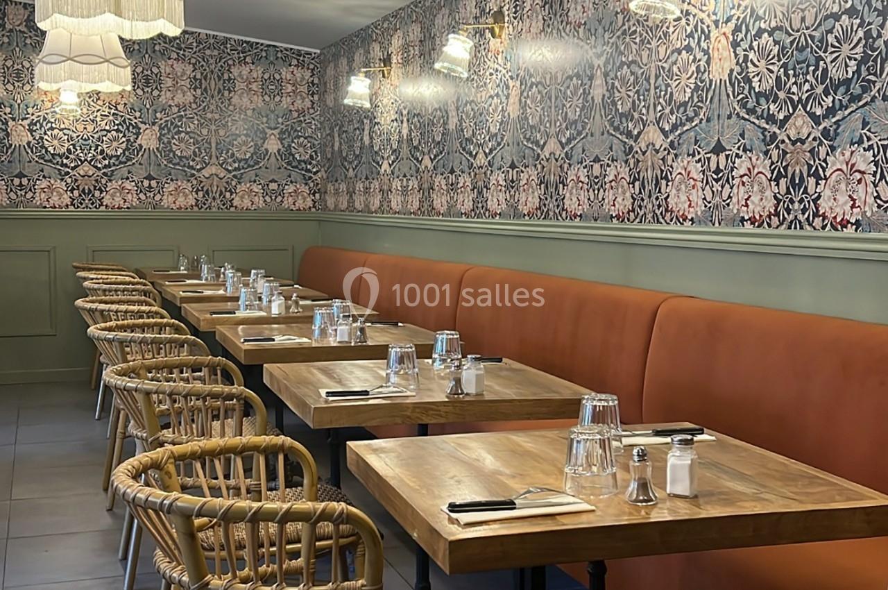 Salle de restaurant avec tables en bois, chaises en rotin, banquette orange et murs ornés de papier peint floral.