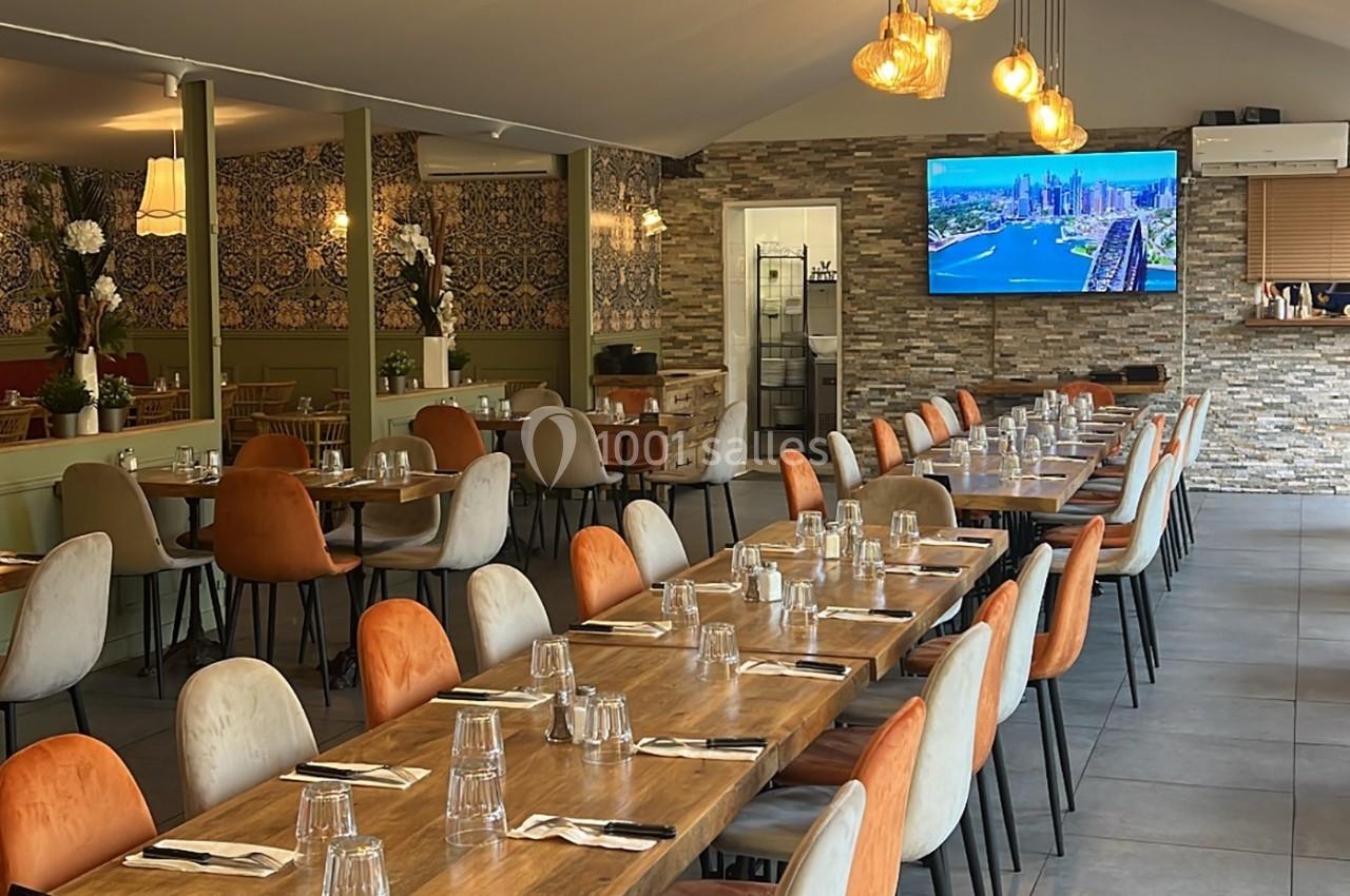 Salle de restaurant moderne avec tables en bois dressées, chaises colorées et écran affichant une vue urbaine.