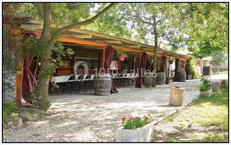 Terrasse extérieure ombragée avec longues tables dressées, entourée de verdure et décorée de tonneaux et plantes.