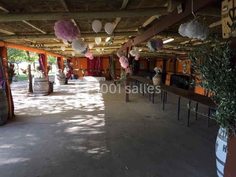 Salle rustique décorée de ballons et de fleurs, avec des tables alignées et des tonneaux en bois, éclairée par la lumière…