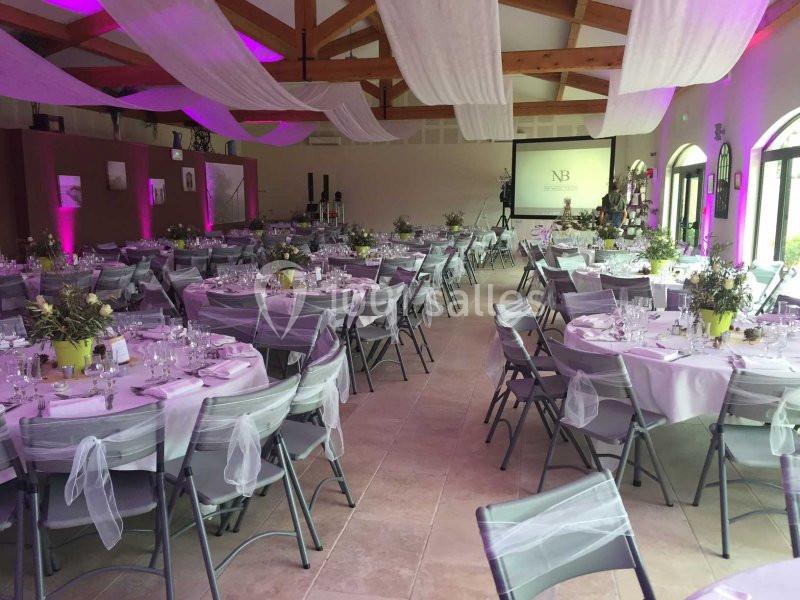 Salle de réception décorée avec des tables rondes, nappes blanches, chaises ornées de rubans et éclairage rose.