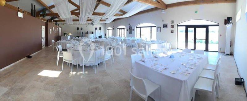 Salle de réception lumineuse avec tables dressées, chaises blanches et poutres apparentes au plafond.