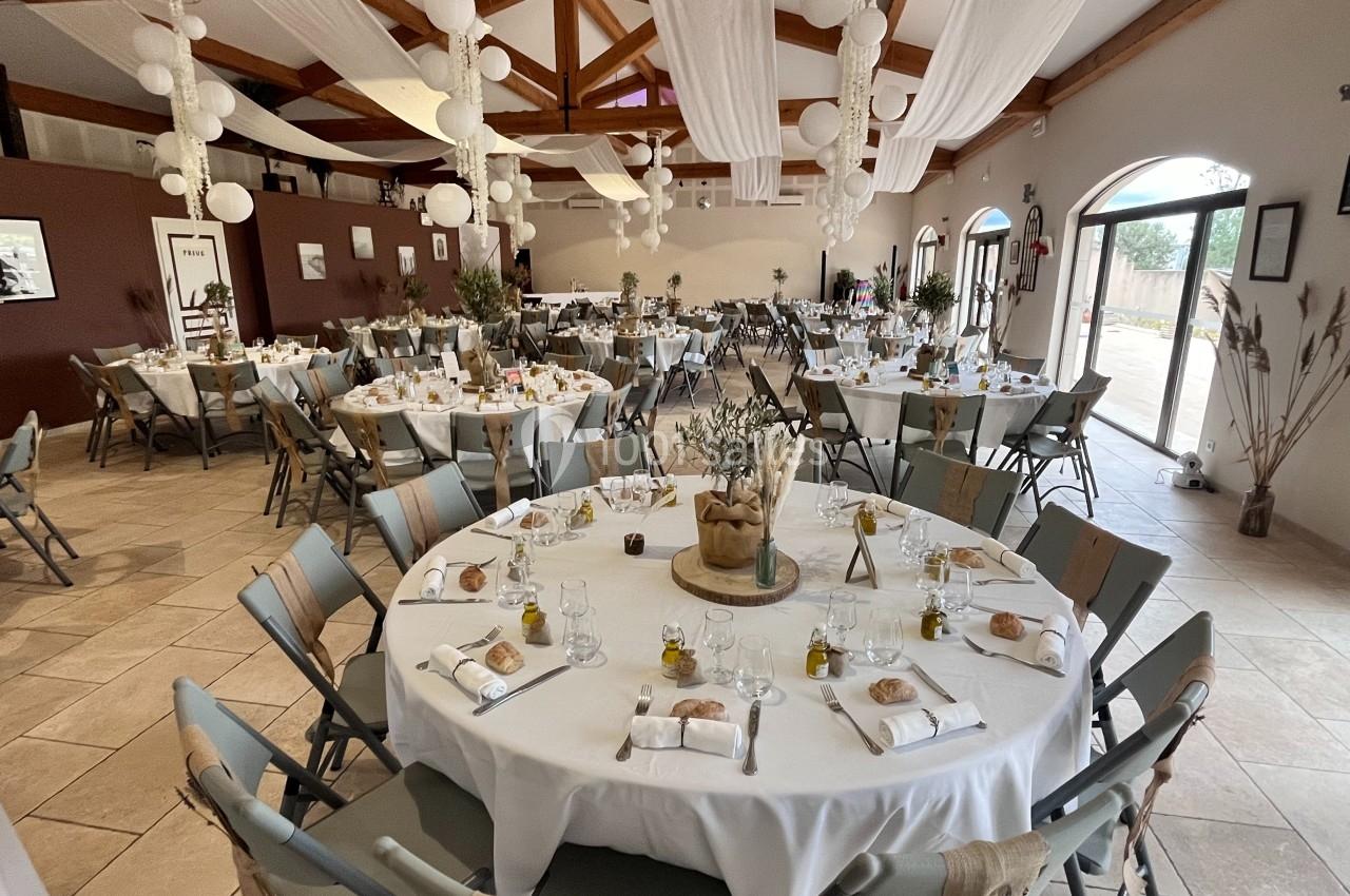 Salle de réception décorée avec des tables rondes dressées, nappes blanches, chaises pliantes et éclairage naturel.