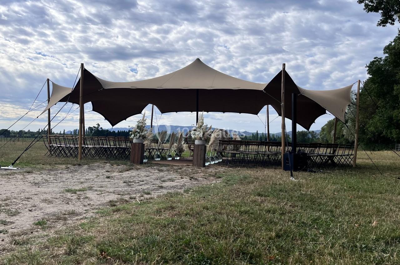 Tente beige montée en extérieur avec des rangées de chaises alignées, entourée de verdure sous un ciel partiellement nuageux.