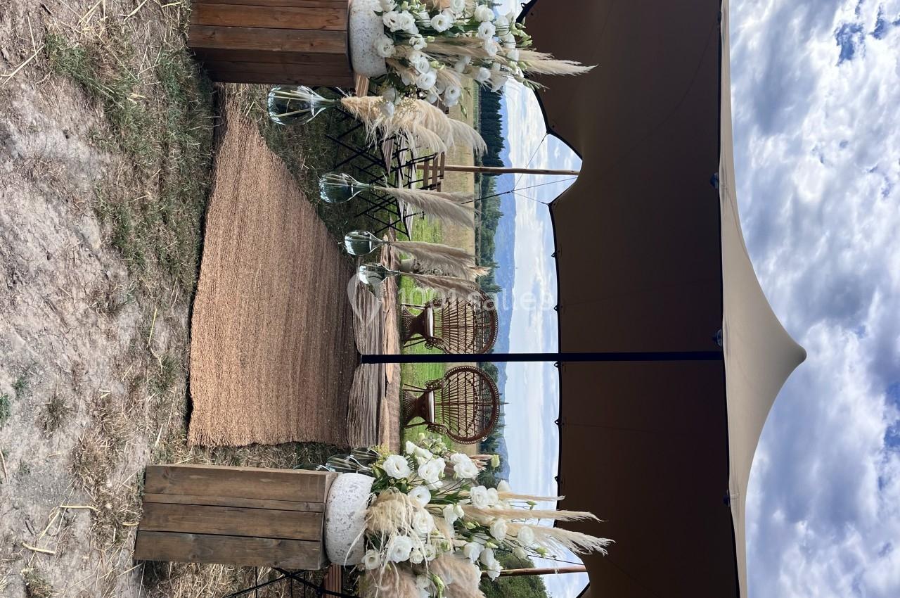 Décoration de cérémonie en extérieur avec tapis, fleurs séchées, chaises en rotin et tente beige sous un ciel nuageux.