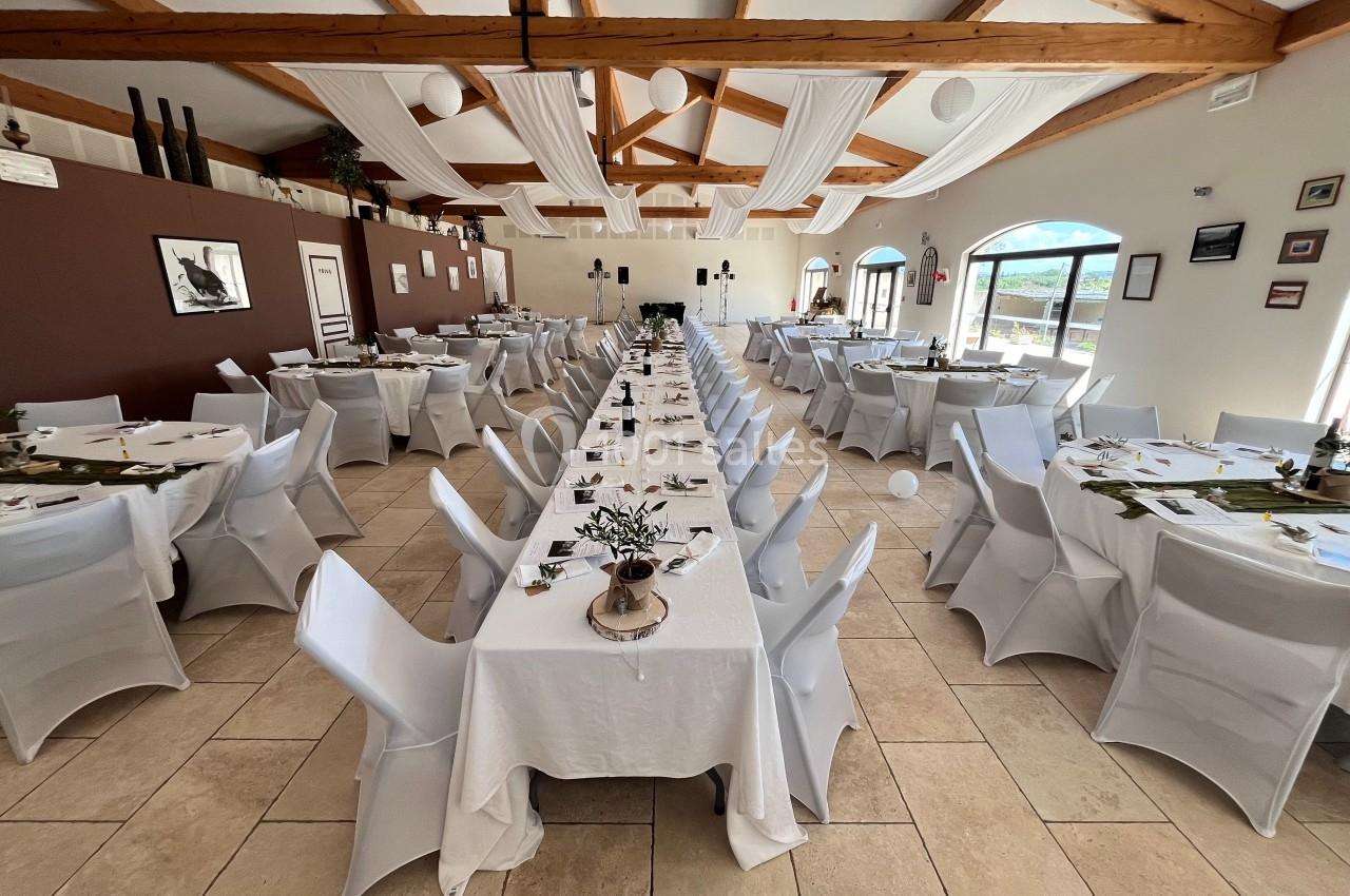 Salle de réception décorée avec des tables alignées, nappes blanches et chaises houssées, sous un plafond en bois.
