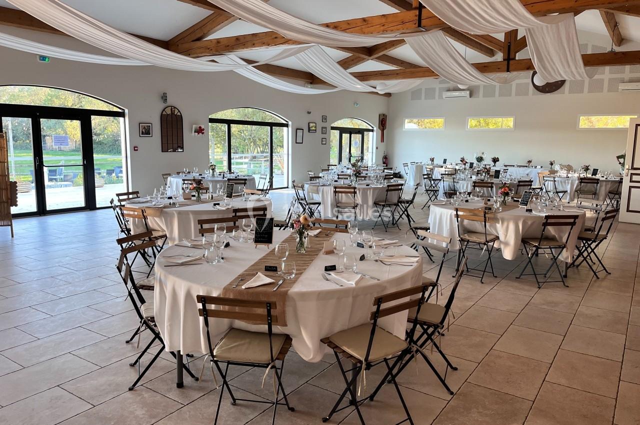 Salle de réception lumineuse avec tables rondes dressées, chaises pliantes et poutres apparentes au plafond.
