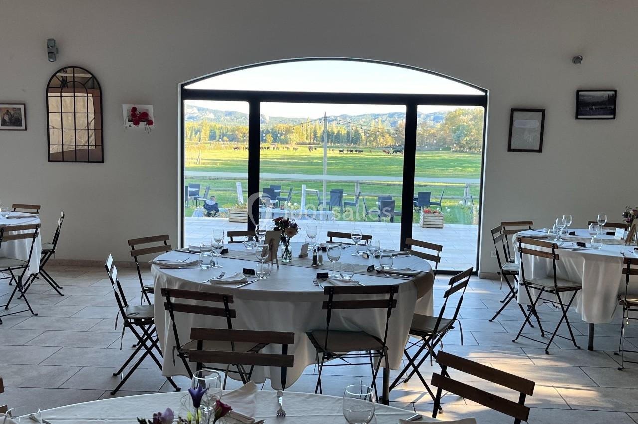Salle de réception lumineuse avec tables dressées, vue sur une terrasse et un paysage verdoyant à travers une grande baie…