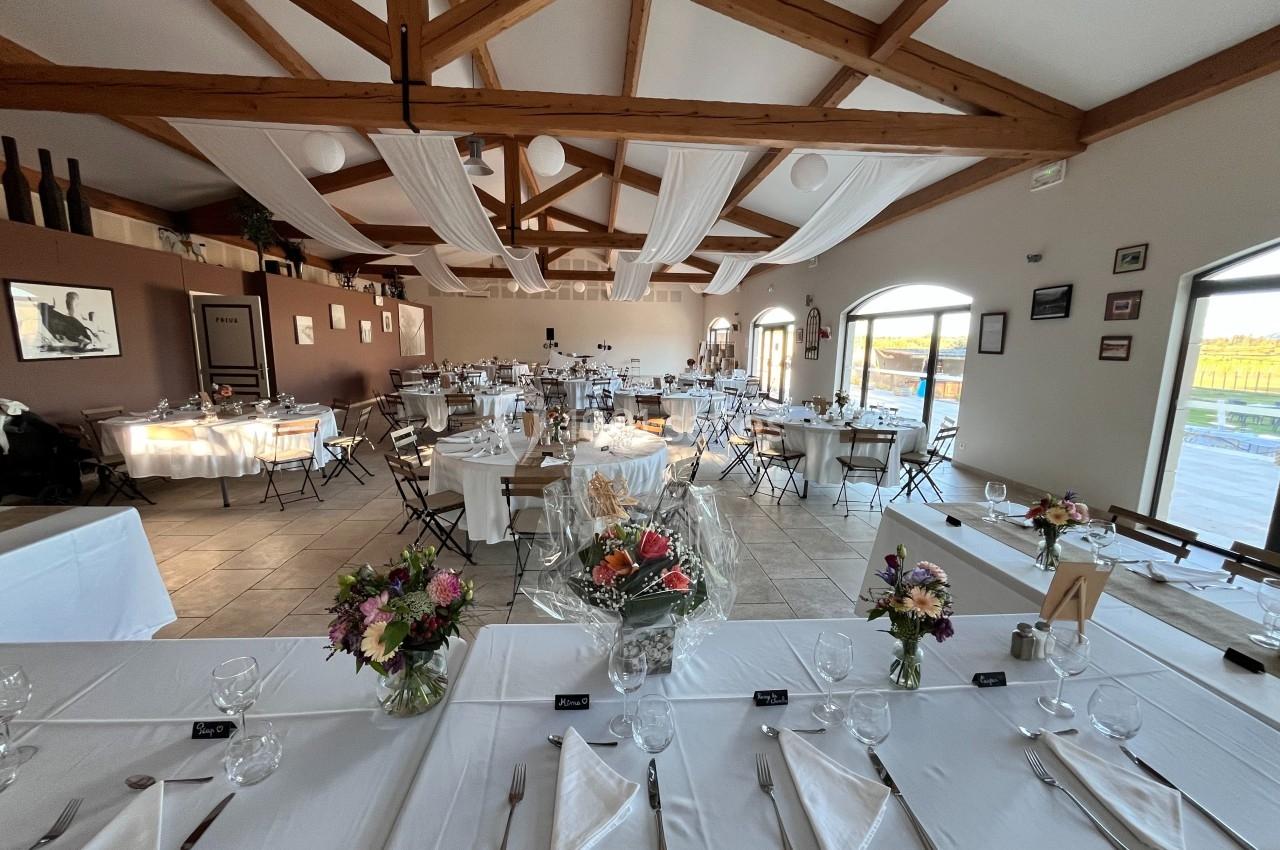 Salle de réception décorée avec des tables dressées, des fleurs et des voilages suspendus sous un plafond en bois.