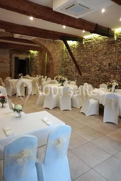 Salle de réception avec murs en pierre, tables rondes et chaises blanches décorées de nœuds beiges.