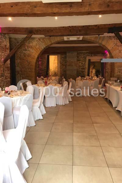 Salle de réception avec murs en pierre, poutres apparentes et tables rondes dressées avec nappes blanches.
