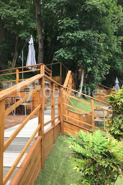Terrasse en bois avec escaliers et garde-corps, entourée de végétation dense et parasols fermés en arrière-plan.