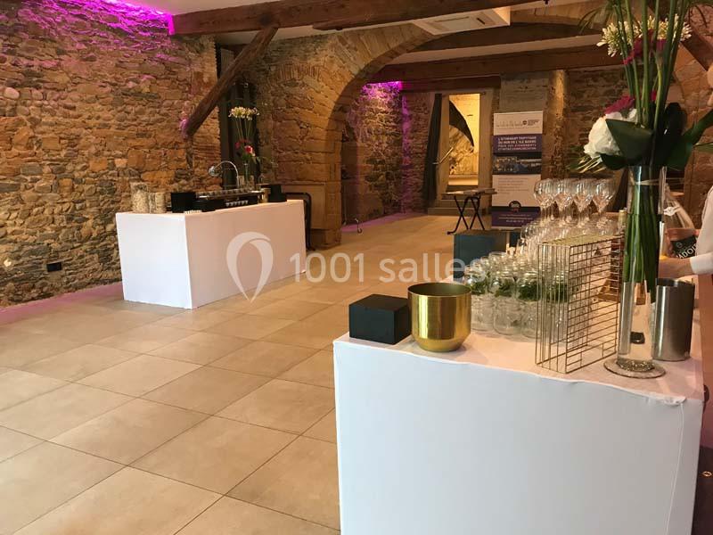 Salle en pierre avec poutres apparentes, décorée pour un événement, tables blanches avec verres et décorations florales.