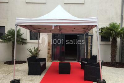 Tente blanche avec mobilier de jardin noir sur tapis rouge, installée devant une entrée vitrée d'un bâtiment beige.
