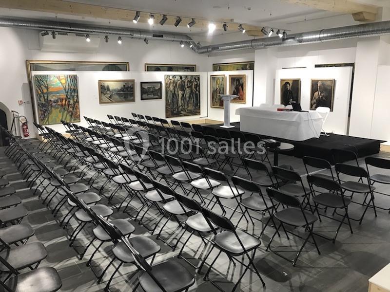 Salle d'exposition avec des rangées de chaises noires alignées devant une scène et des peintures accrochées aux murs.