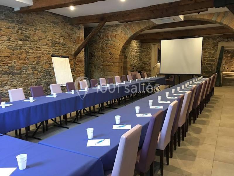 Salle de réunion avec longues tables couvertes de nappes bleues, chaises alignées, écran de projection et paperboard.