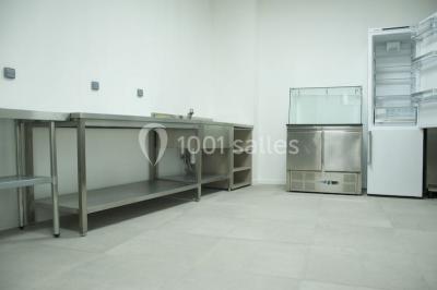 Cuisine professionnelle vide avec plans de travail en inox, évier, étagères ouvertes et réfrigérateurs en arrière-plan.