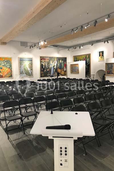 Salle de conférence avec des rangées de chaises noires vides, un pupitre et un micro au premier plan.