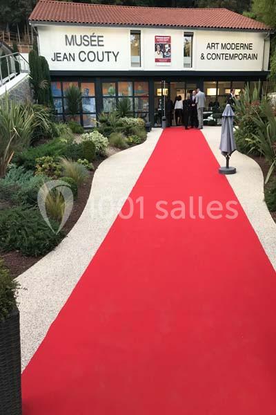 Entrée du musée Jean Couty avec un tapis rouge, entourée de végétation et de visiteurs à l'arrière-plan.
