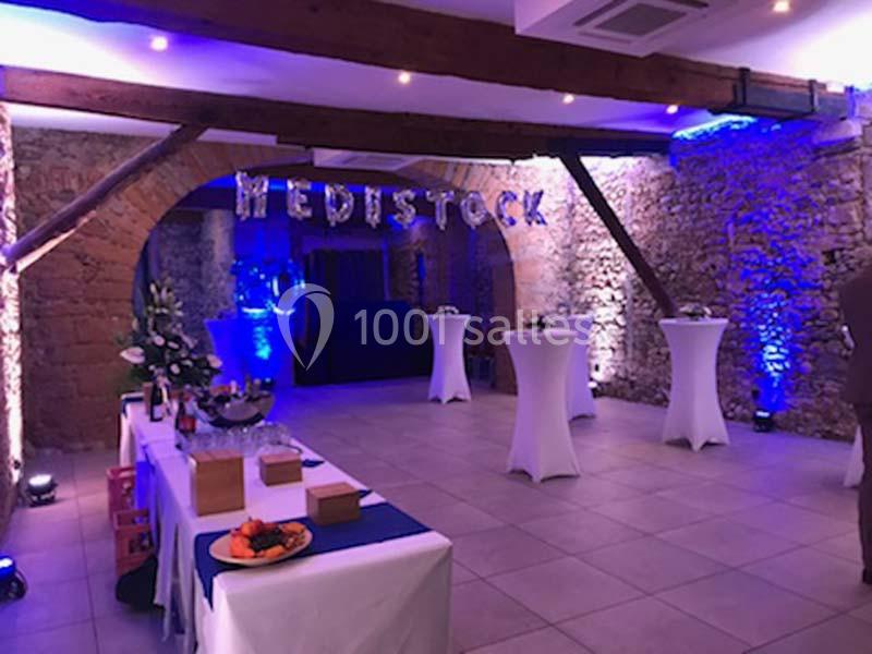 Salle de réception décorée avec des lumières bleues, des tables hautes et un buffet, dans un cadre en pierre apparente.