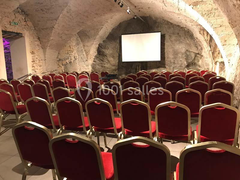 Salle voûtée en pierre avec des rangées de chaises rouges disposées face à un écran de projection blanc.