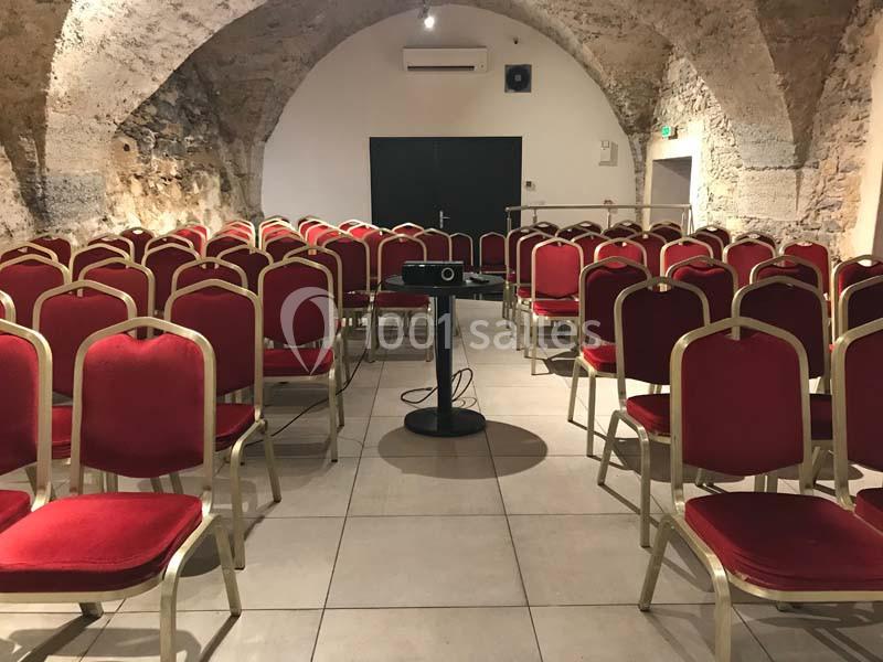 Salle voûtée en pierre avec des rangées de chaises rouges alignées face à un projecteur sur pied.