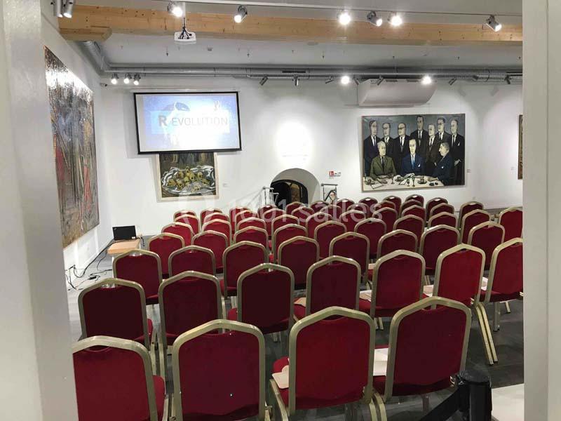 Salle de conférence avec des rangées de chaises rouges, un écran de projection et des tableaux accrochés aux murs.