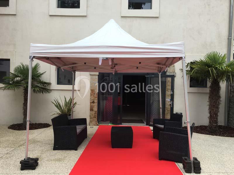 Tente blanche avec salon de jardin noir sur tapis rouge devant une entrée vitrée, entourée de palmiers.