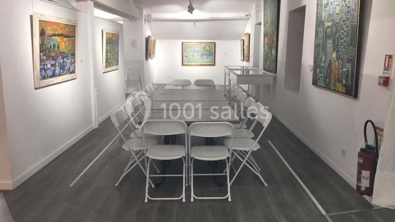 Salle d'exposition lumineuse avec une grande table entourée de chaises pliantes, entourée de peintures accrochées aux murs.