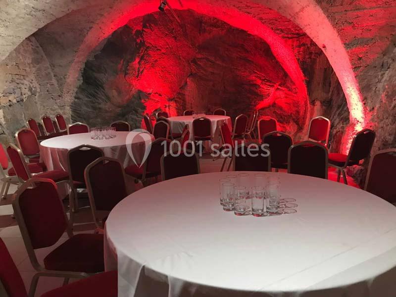 Salle voûtée en pierre éclairée de rouge, aménagée avec des tables rondes dressées de nappes blanches et des chaises rouges.