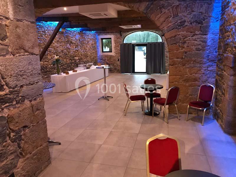 Salle en pierre avec plafond en poutres apparentes, tables et chaises rouges, buffet dressé et éclairage bleu.