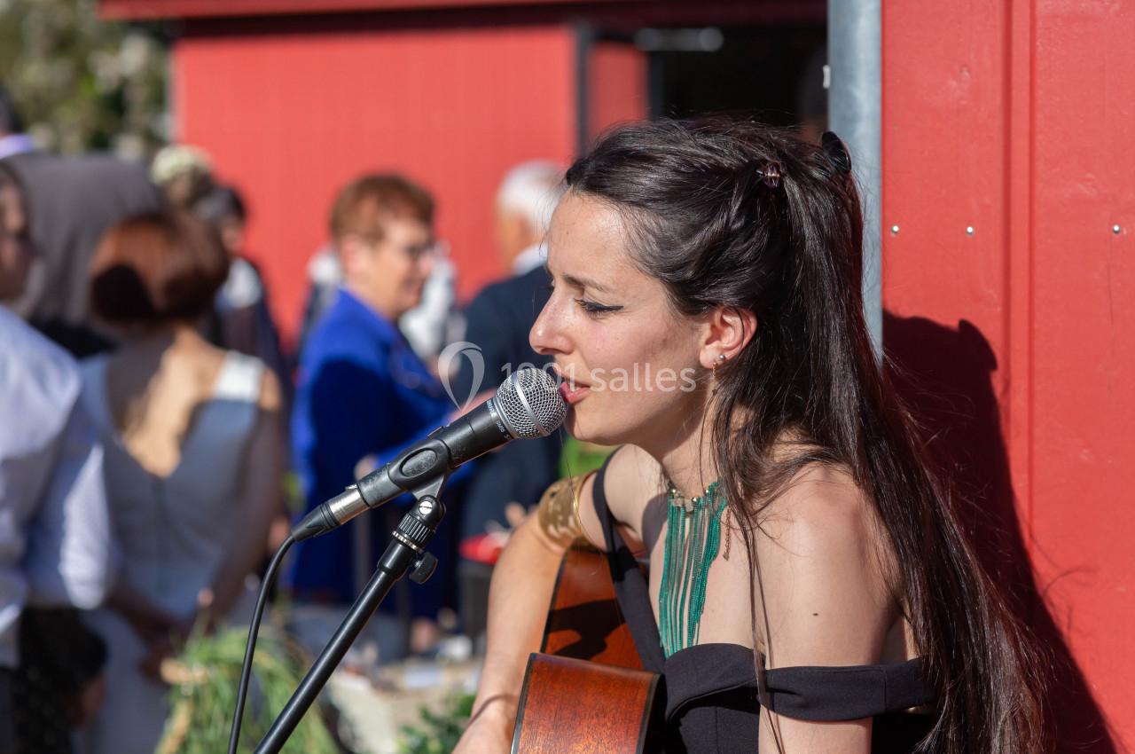 Une femme chante en jouant de la guitare devant un micro, avec des personnes en arrière-plan lors d'un événement extérieur.