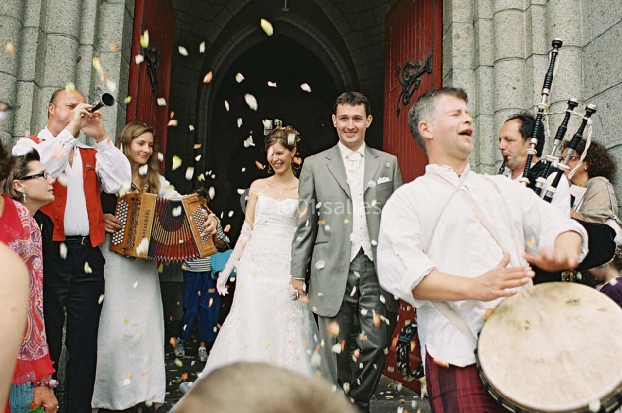 Un couple de mariés sort d'une église sous une pluie de confettis, entouré de musiciens jouant des instruments traditionnels.