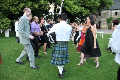 Un couple de mariés ému écoute un joueur de cornemuse, entouré d'invités applaudissant dans une salle lumineuse.