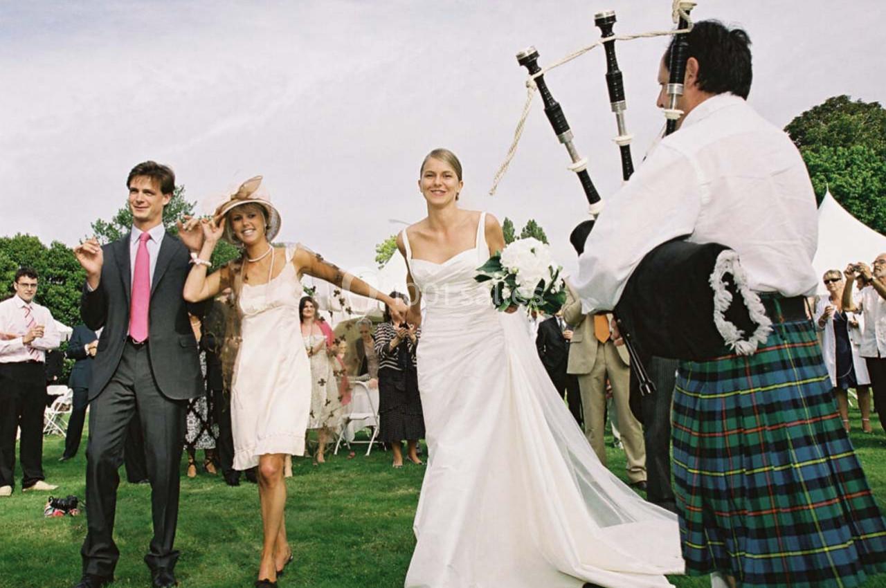 Un joueur de cornemuse en kilt joue lors d'un mariage en plein air, avec des invités dansant sur une pelouse.