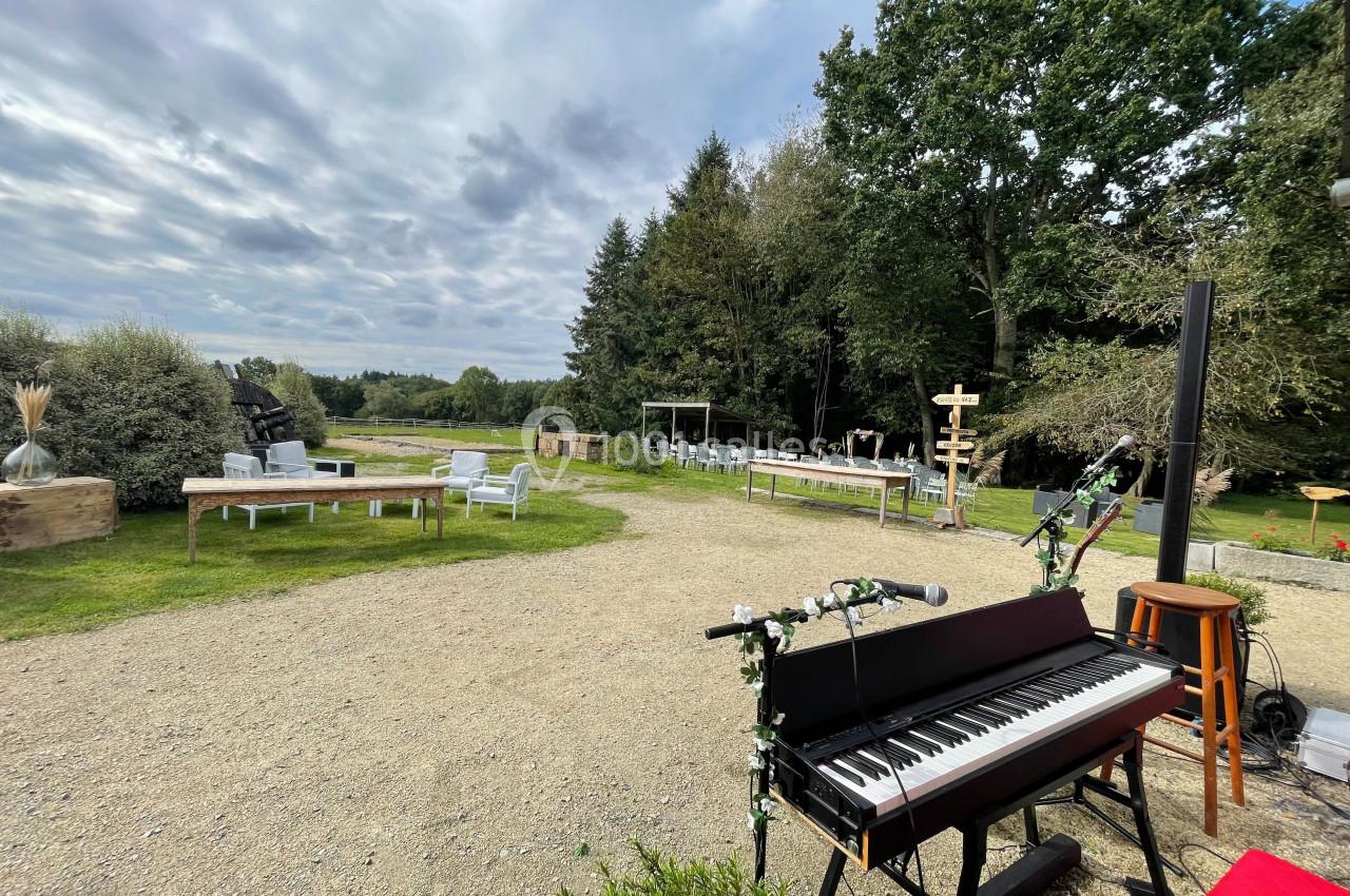 Piano décoré en extérieur sur une terrasse en gravier, entouré de mobilier de jardin et d'arbres sous un ciel nuageux.