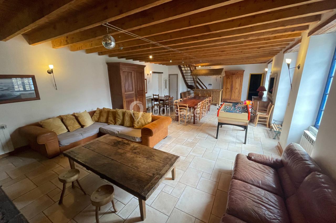 Salle de séjour spacieuse avec poutres apparentes, canapés, table en bois, baby-foot et grande table à manger.