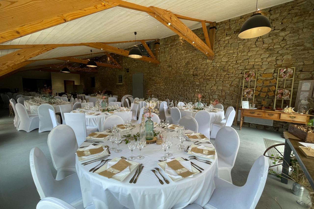 Salle de réception décorée avec des tables rondes dressées, nappes blanches, chaises houssées et murs en pierre apparente.