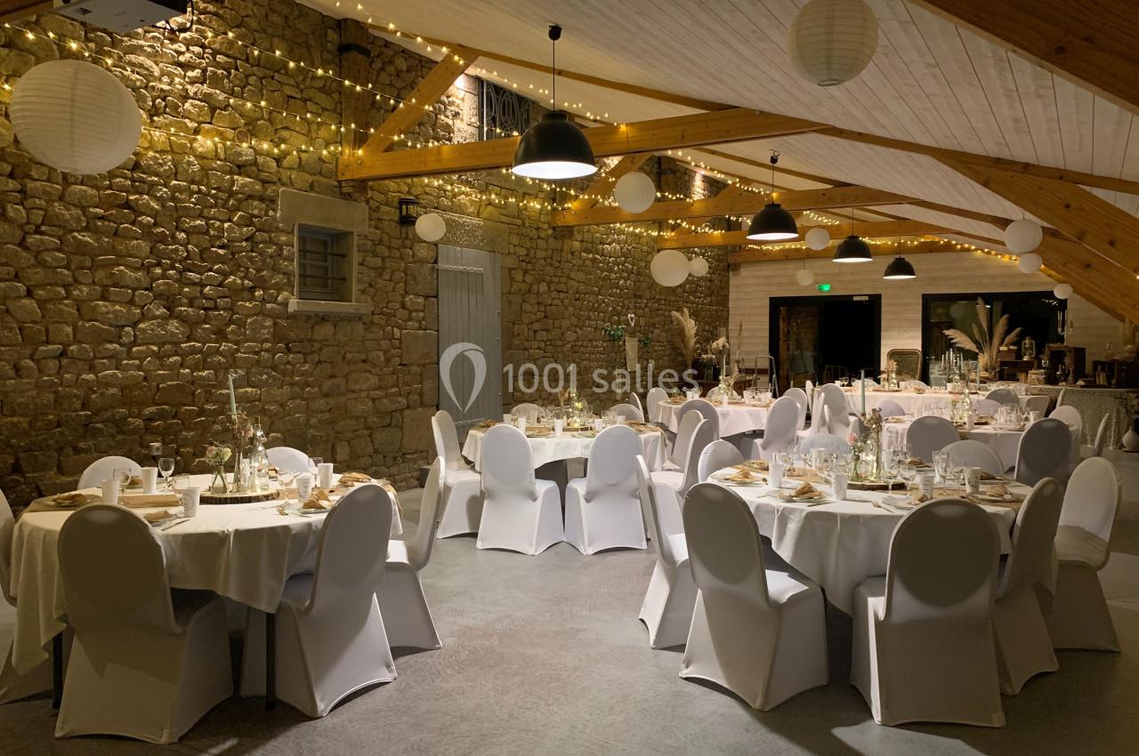 Salle de réception décorée avec des tables rondes dressées, des chaises blanches et des guirlandes lumineuses suspendues.