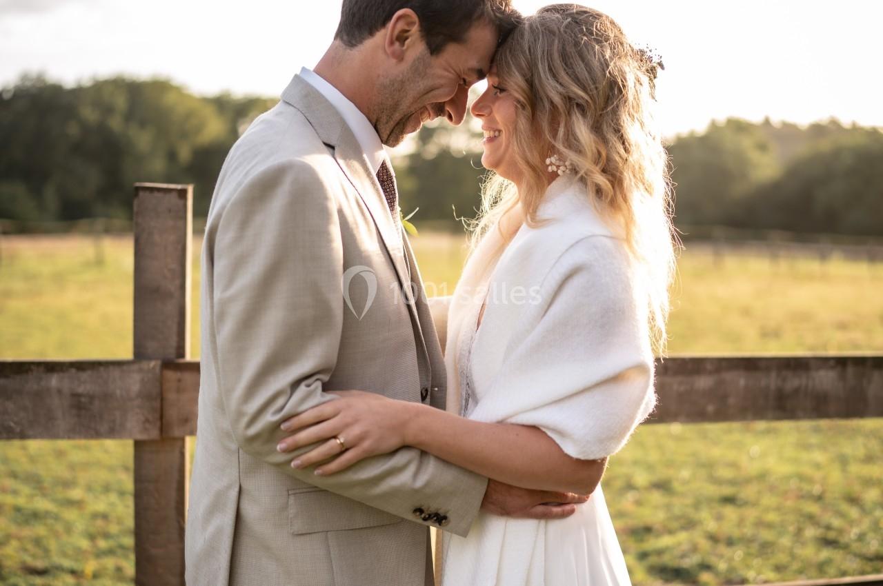 Un couple souriant s'enlace tendrement devant une clôture en bois, sous une lumière douce au coucher du soleil.