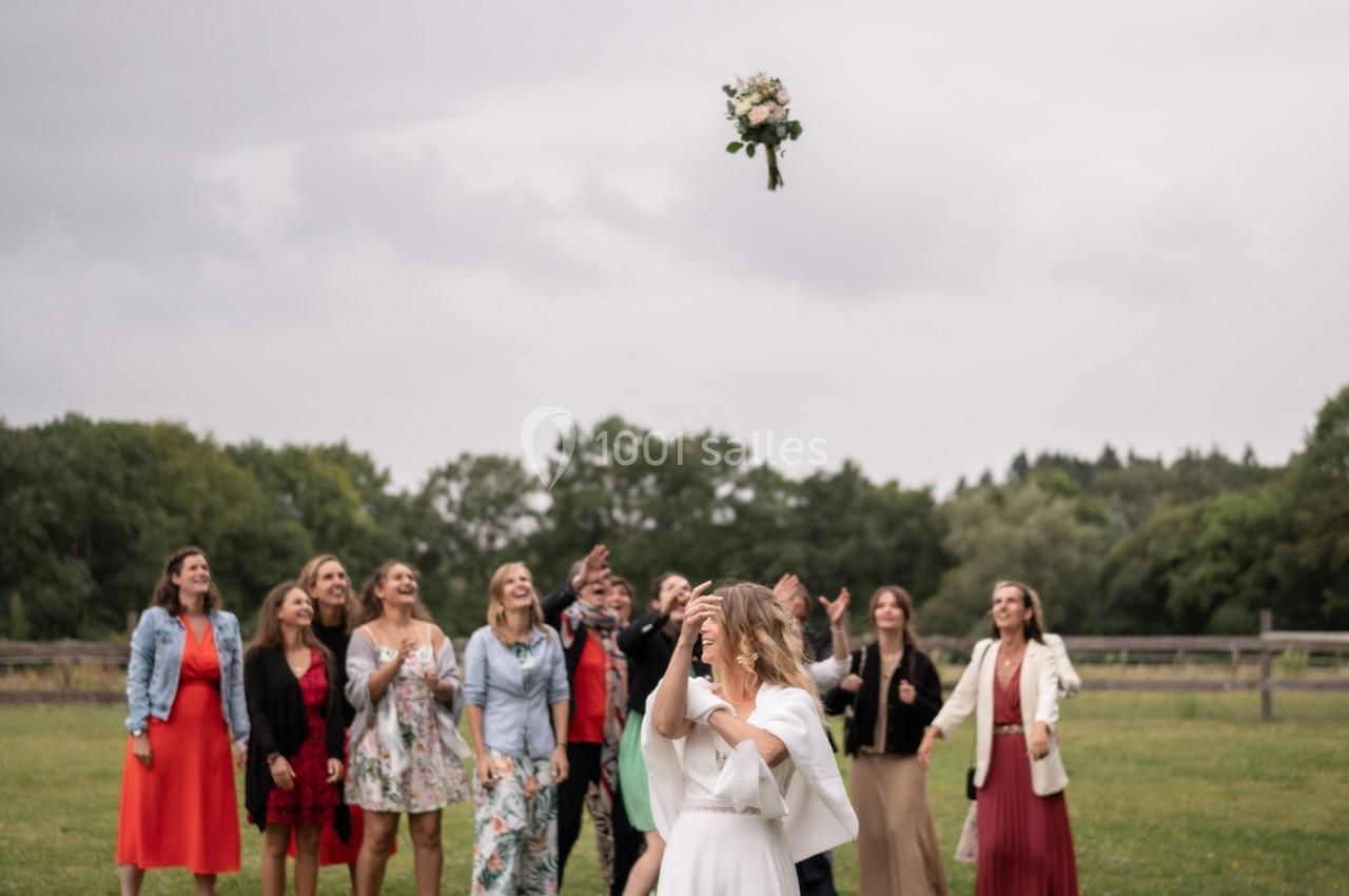 Une mariée lance son bouquet de fleurs vers un groupe de femmes dans un champ sous un ciel nuageux.