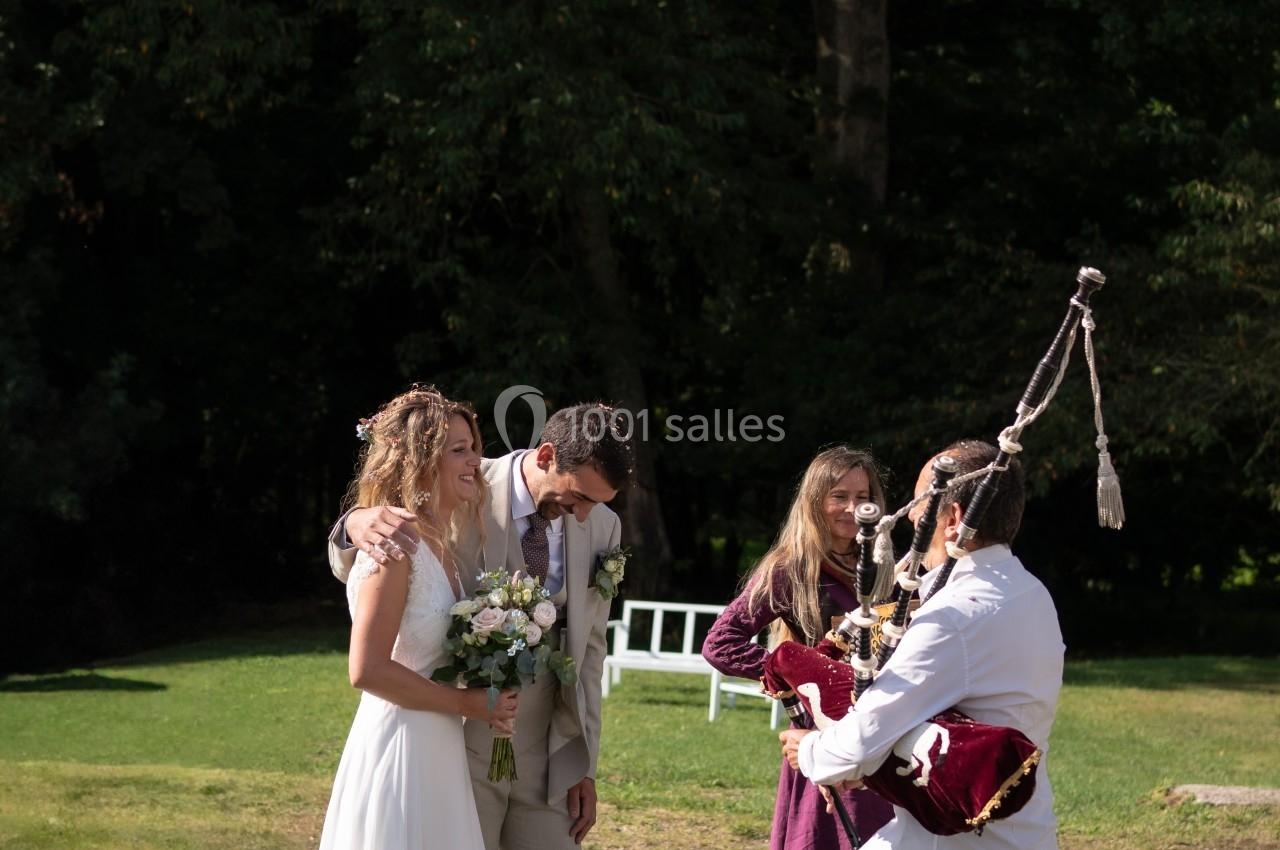Un couple de mariés sourit en extérieur, accompagné de musiciens jouant de la cornemuse dans un cadre verdoyant.