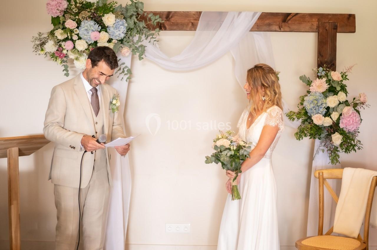 Un couple échange des vœux devant une arche décorée de fleurs lors d'une cérémonie de mariage en intérieur.