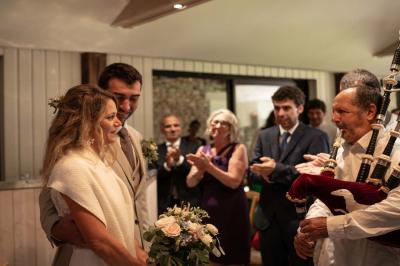 Un couple de mariés ému écoute un joueur de cornemuse, entouré d'invités applaudissant dans une salle lumineuse.