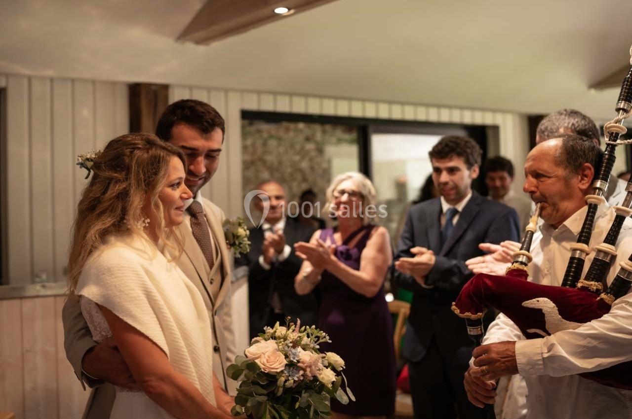 Un couple de mariés ému écoute un joueur de cornemuse, entouré d'invités applaudissant dans une salle lumineuse.